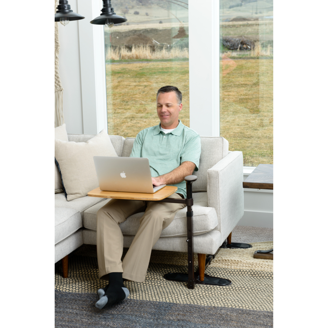 A man in a light green polo and beige pants uses his laptop on the Stander Omni Tray – TV Tray & Standing Aid while seated on a light sofa. Large windows behind him showcase a grassy outdoor view.