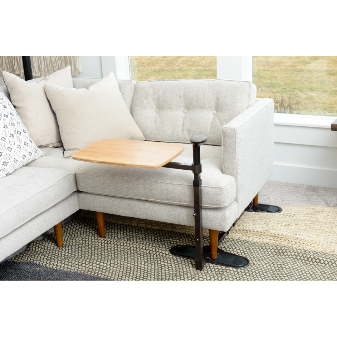 A Stander Omni Tray – TV Tray & Standing Aid, featuring a wooden swivel table and black adjustable support arm, is attached to a light-colored sectional sofa with beige cushions in a bright living room.