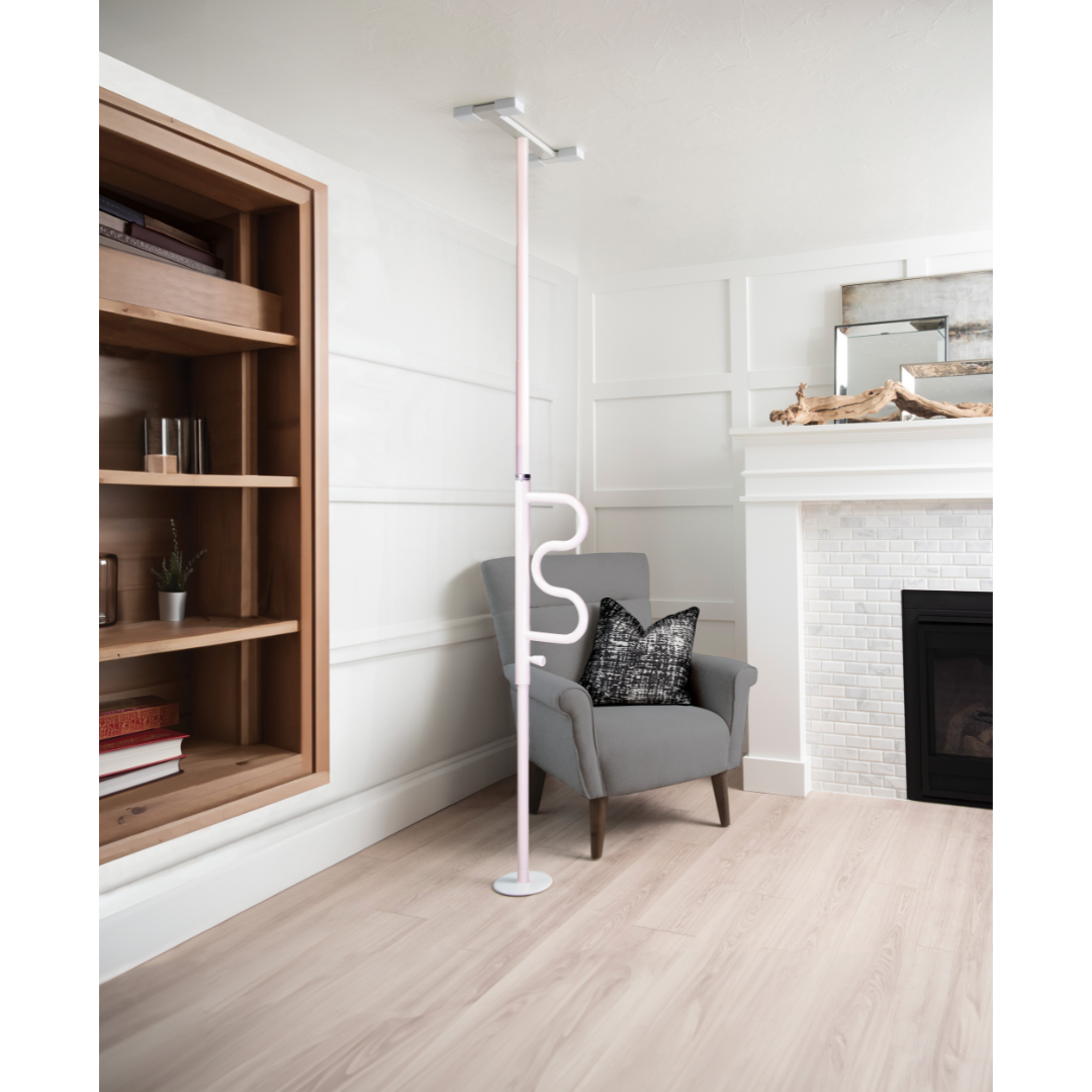 A Stander Wonder Pole - Fall Prevention Safety Pole For The Home extends from floor to ceiling beside a gray armchair in a bright living room with wooden floors, built-in shelves, and a white fireplace, providing convenient sit-to-stand support.