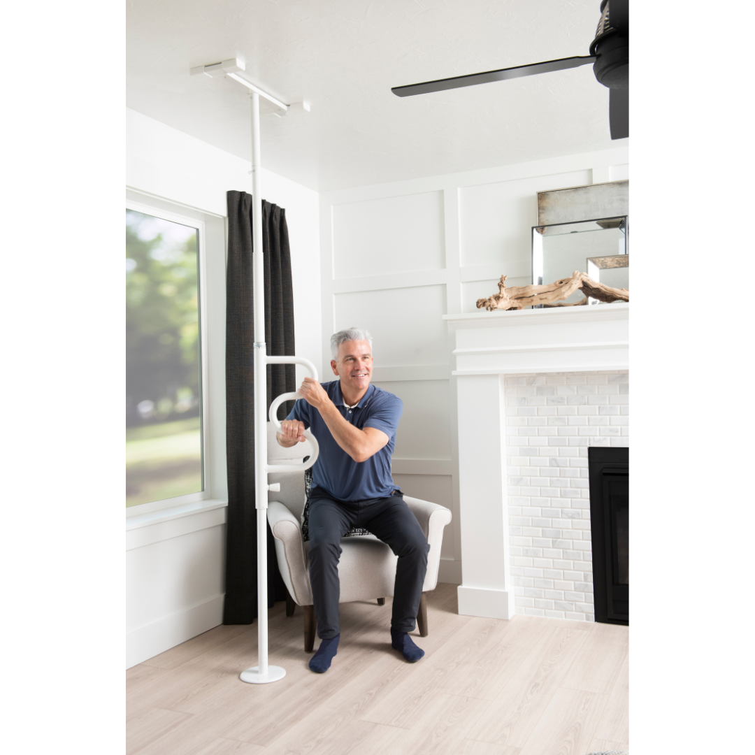 An older man sits on a chair in a living room, holding the Stander Wonder Pole - Fall Prevention Safety Pole for stability. The room features a large window, black curtains, a fireplace, and light wood floors.