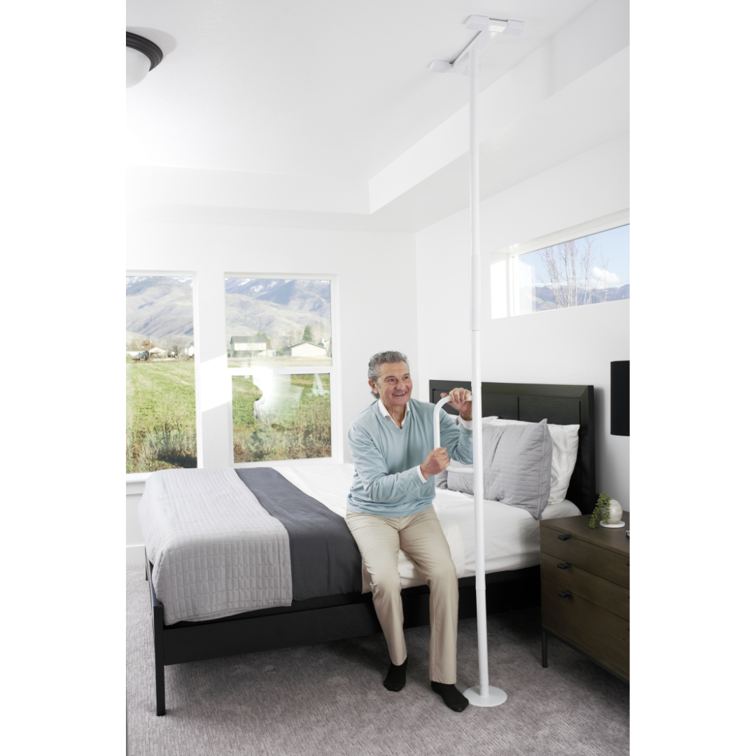 An older adult holds the Stander Wonder Pole Lite—Fall Prevention Safety Pole For The Home—beside a bed in a bright bedroom with large windows and a scenic view, highlighting its use for fall prevention.