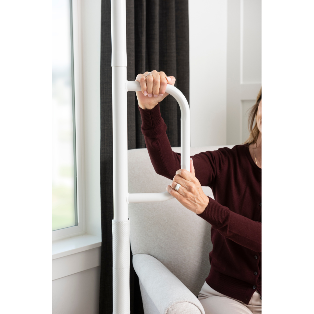 A woman in a maroon sweater sits in a chair, holding the Stander Wonder Pole Lite—Fall Prevention Safety Pole for the Home—in a bright room with a window and dark curtains.