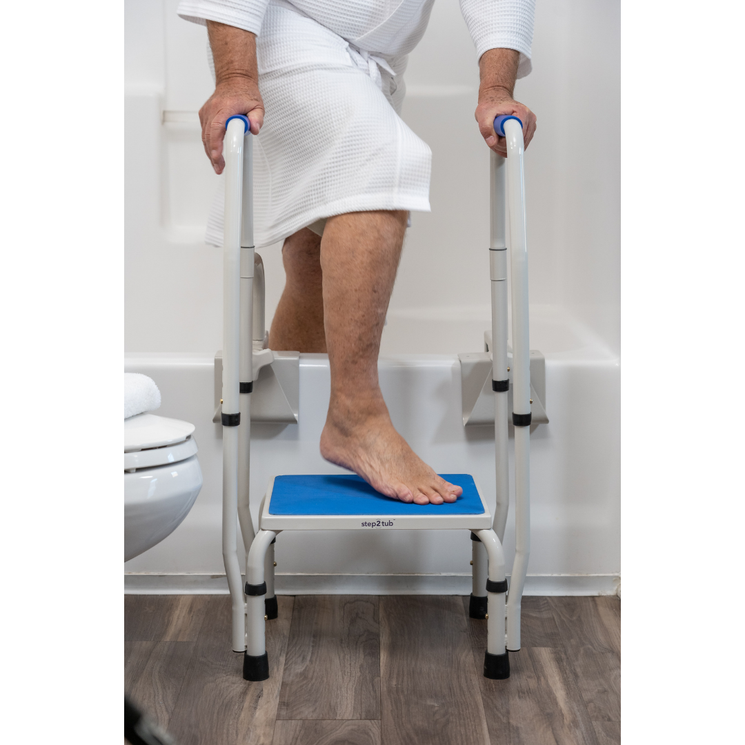 Wearing a white robe, a person uses the Step2Tub Bathtub Step Stool with handrails to safely step into a bathtub in a bathroom featuring wood-patterned flooring, enhancing fall prevention.