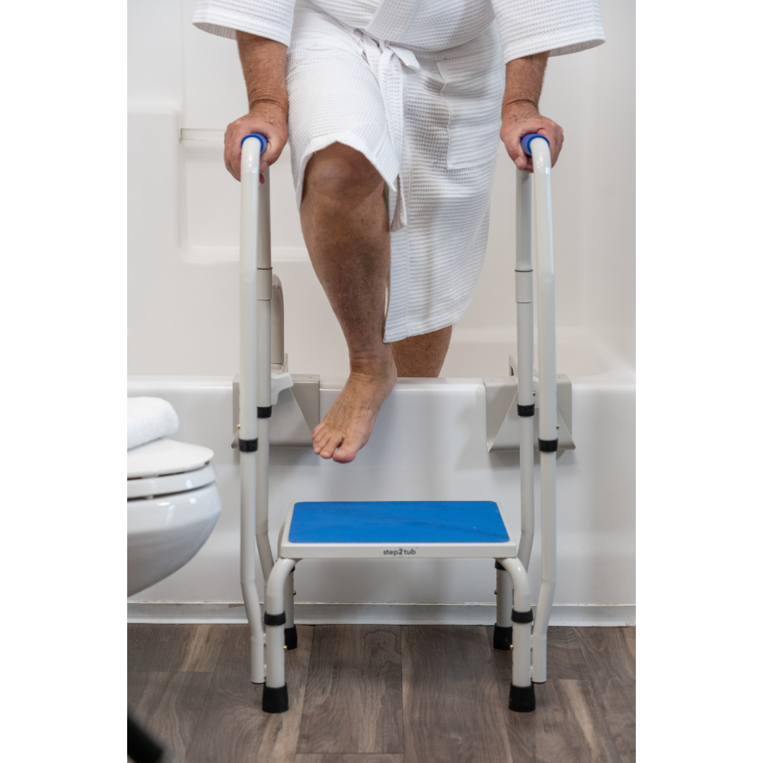 A person in a white robe uses the Step2Tub Bathtub Step Stool with handrails to safely step into or out of a bathtub in a bathroom with wood flooring, enhancing fall prevention.