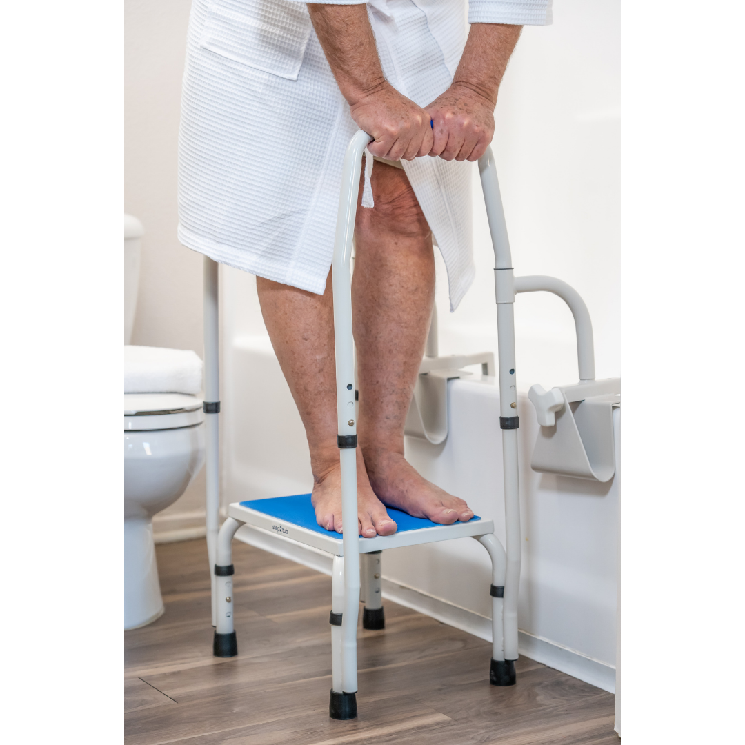An older adult in a white bathrobe uses the Step2Tub Bathtub Step Stool with handrails for support in the bathroom. A toilet and wall-mounted grab bar are visible, underscoring safety and fall prevention measures.
