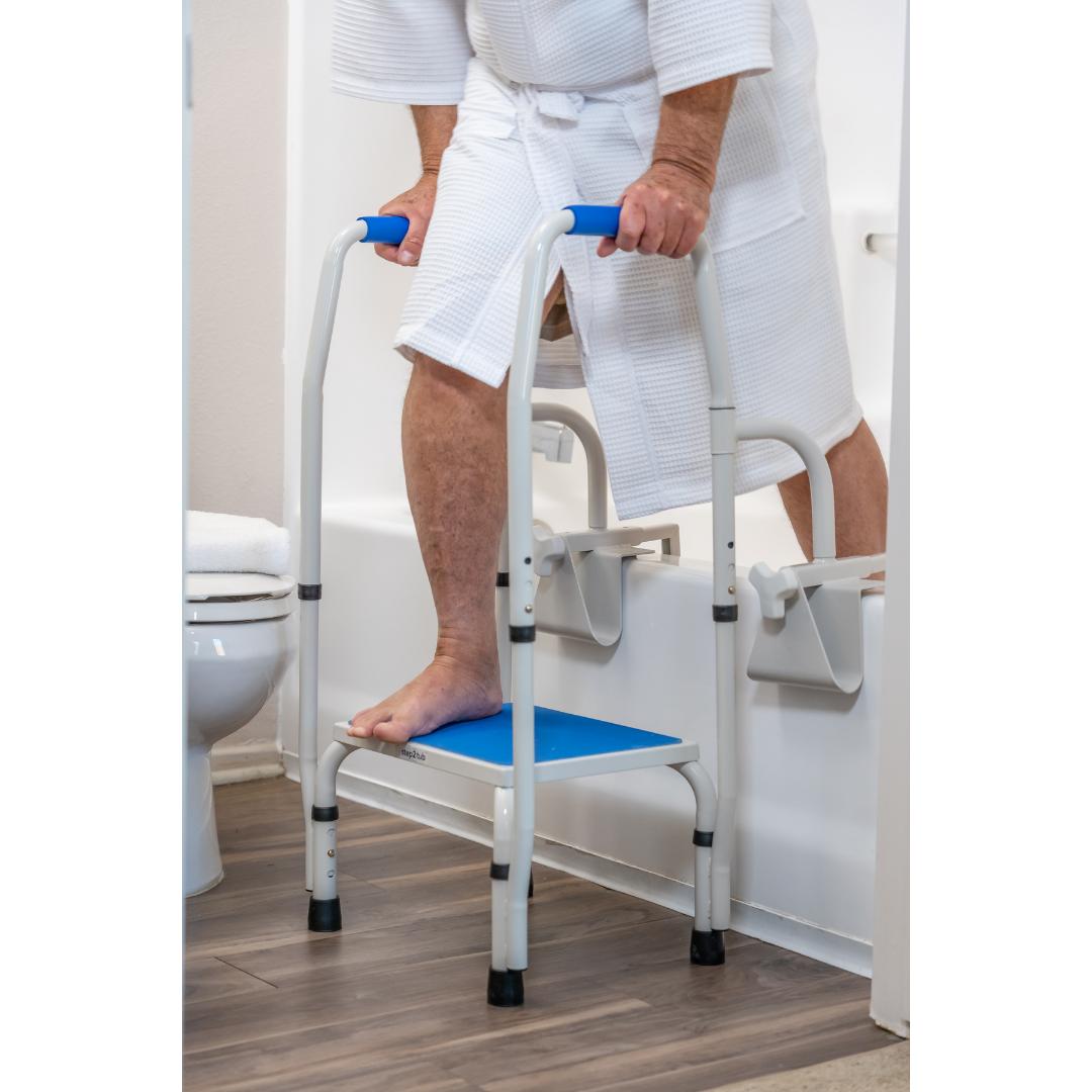 Wearing a white bathrobe, a person uses the Step2Tub Bathtub Step Stool with handles to safely step into the tub in a bathroom with wood floors and a nearby toilet, enhancing safety and fall prevention.