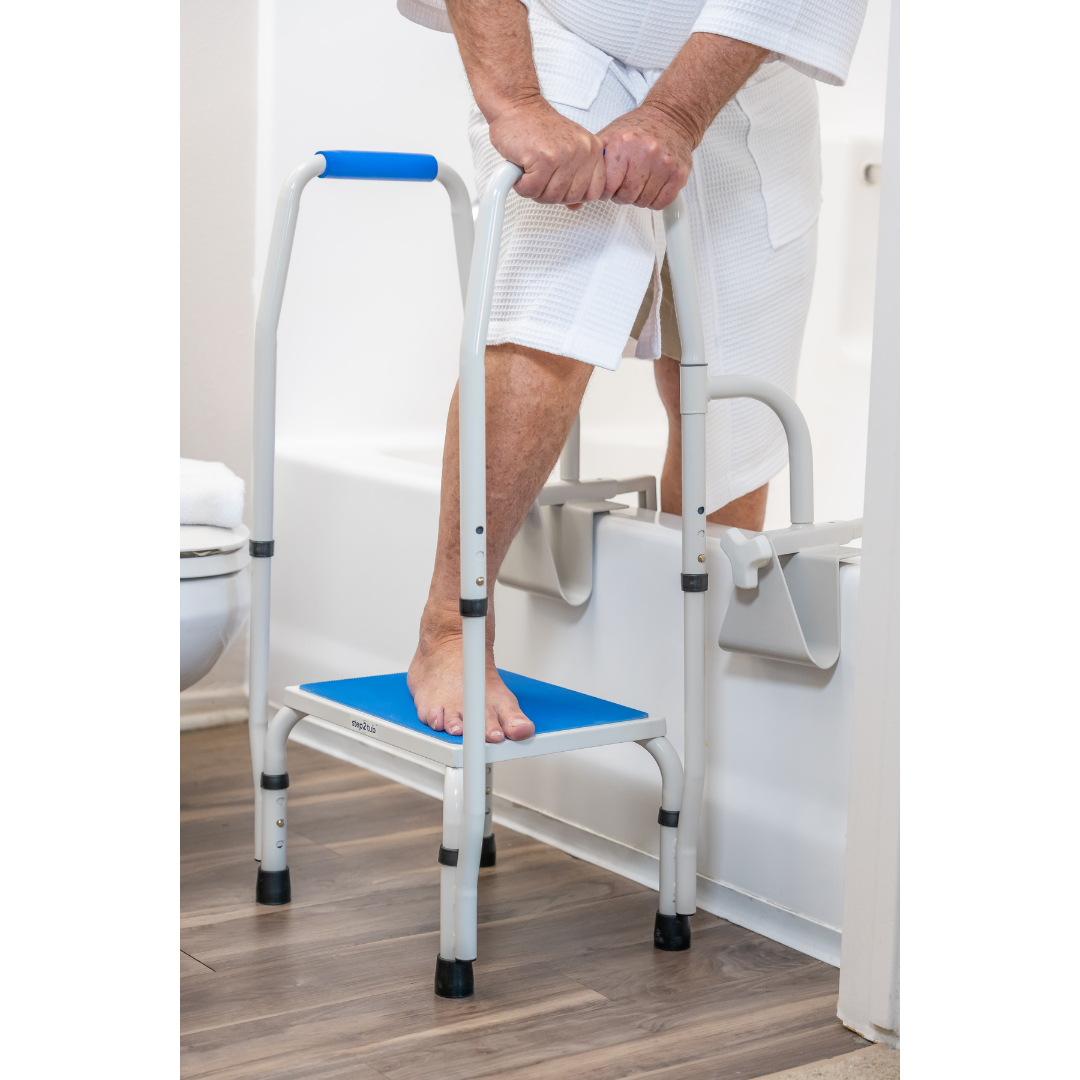 An older adult in a white robe uses the Step2Tub Bathtub Step Stool with handrail to safely enter a bathtub in a bathroom with wooden flooring, helping prevent falls.