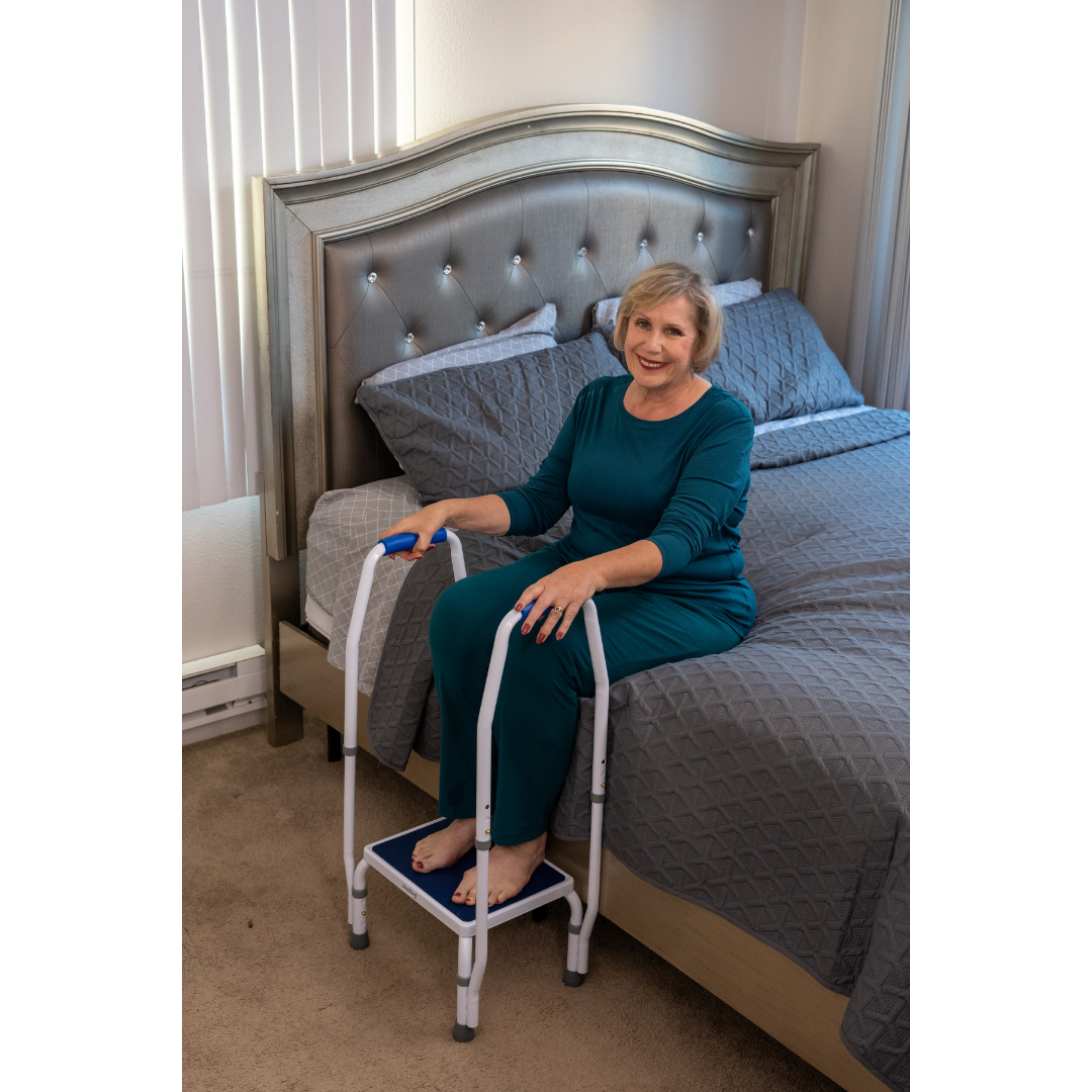 An older woman in teal pajamas smiles while sitting on her bed, holding a Step2Bed Mini Bedside Stepper with handrails—a helpful fall prevention aid—in her bright bedroom with gray bedding and a tufted headboard.