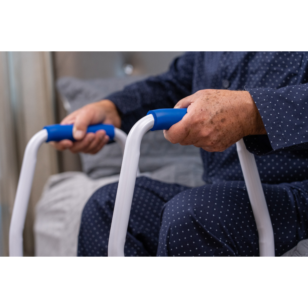 A person in navy blue polka dot pajamas sits by a bed, holding the handles of a white and blue Step2Bed Mini Bedside Stepper, a mobility aid designed for fall prevention.