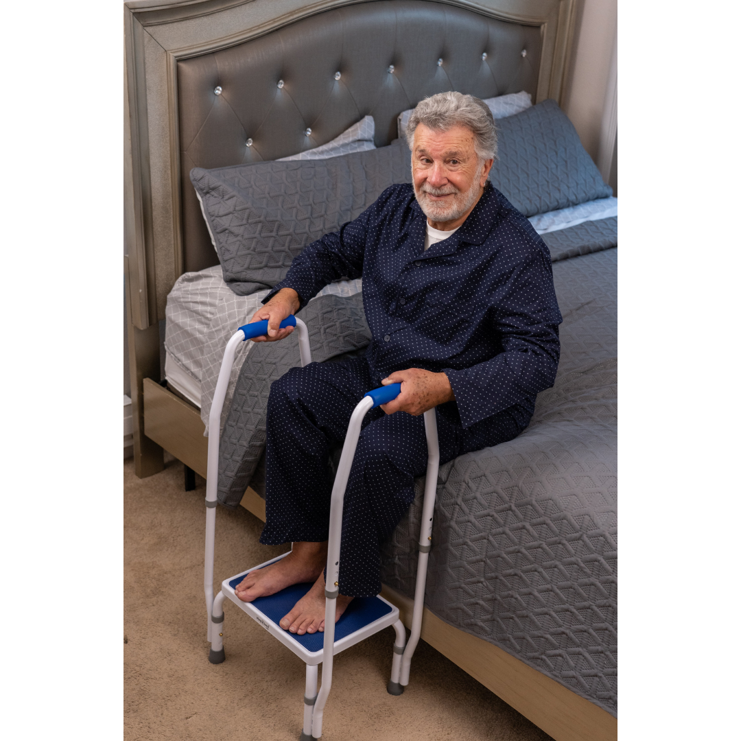 An older man in dark pajamas uses the Step2Bed Mini Bedside Stepper, a white and blue mobility aid, to stand up from the edge of a neatly made bed, smiling as he holds the handles for support to promote safety and prevent falls.