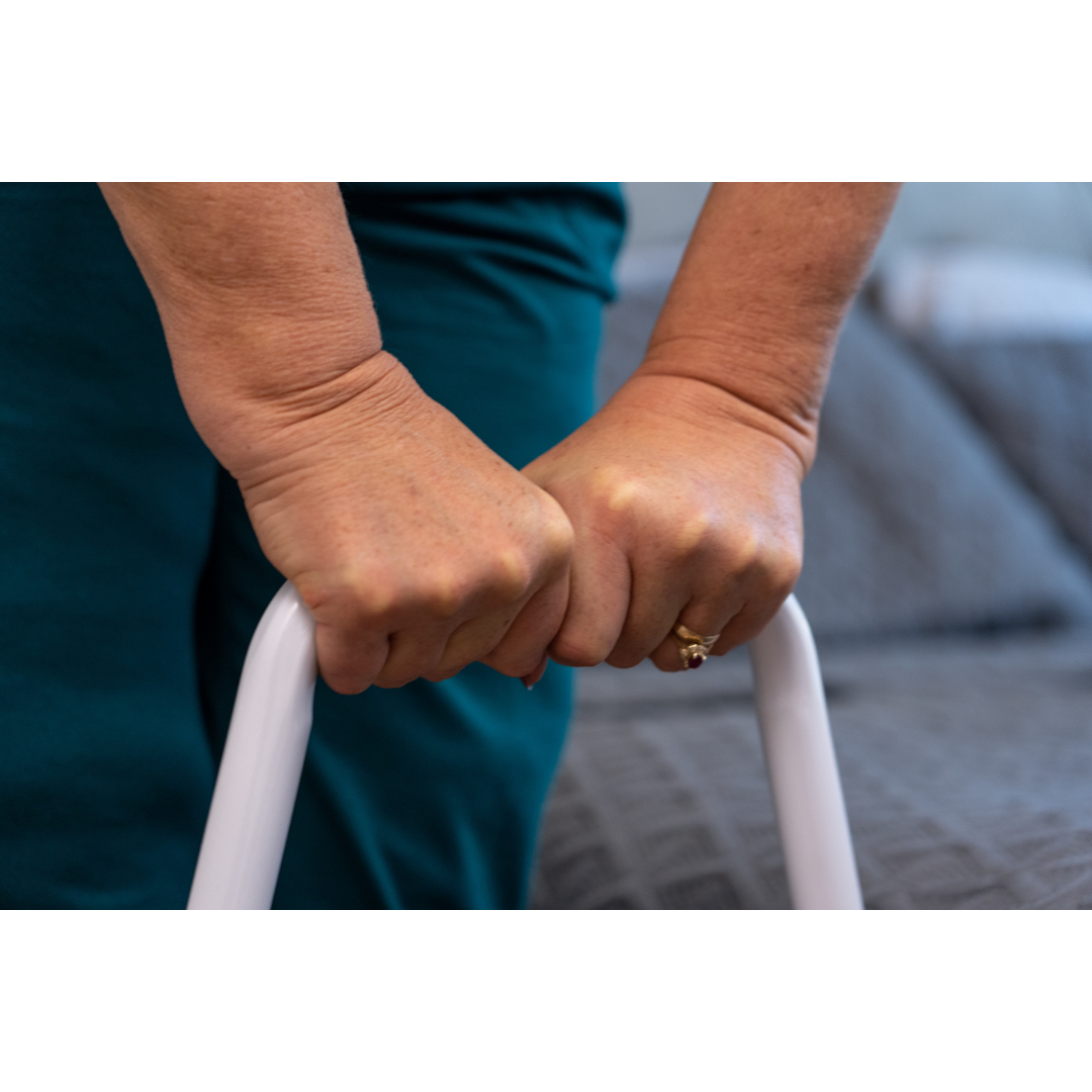 Close-up of an older adult’s hands gripping a white Step2Bed Mini Bedside Stepper rail, with a bed and grey blanket in the background. The person is wearing a gold ring and a teal outfit.