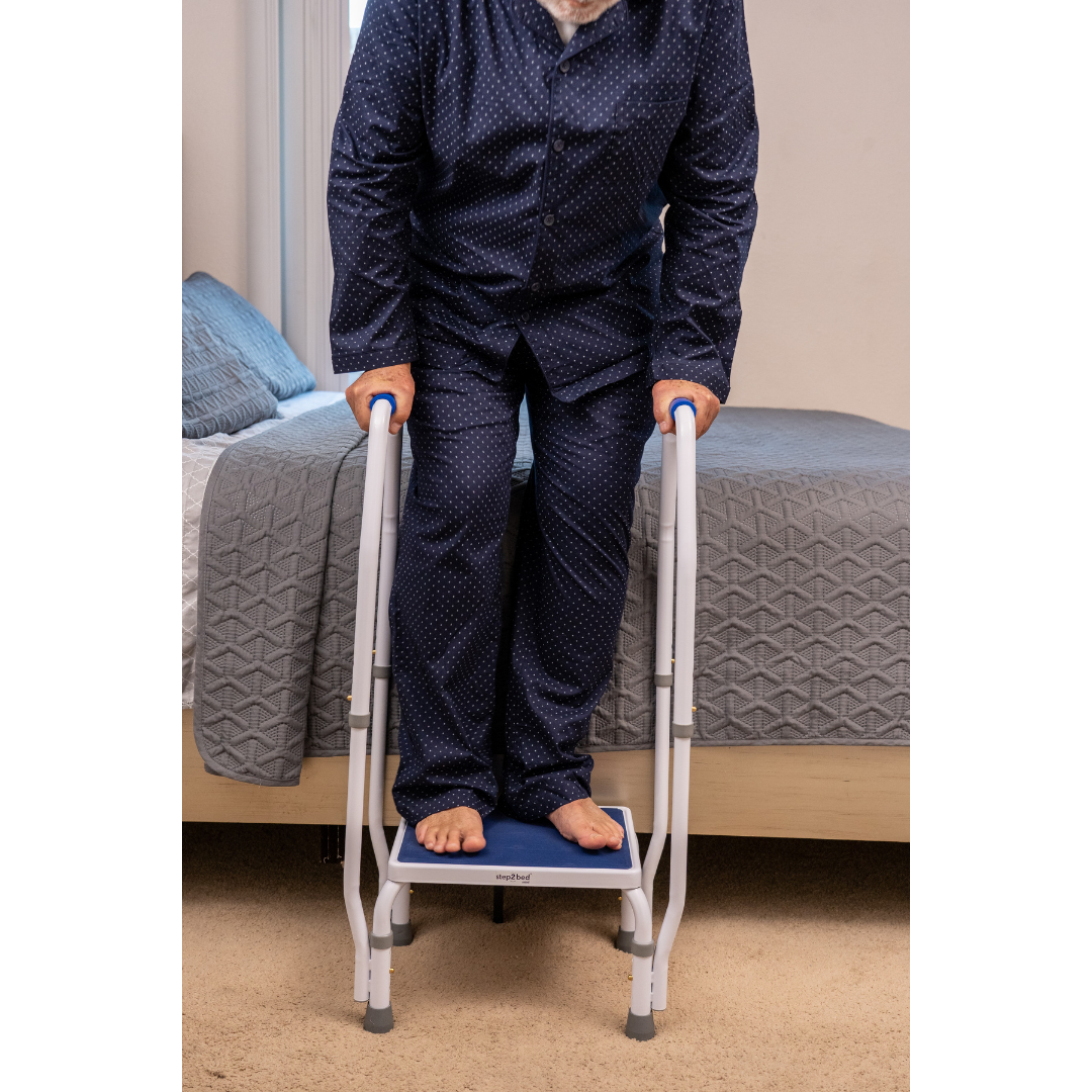 An older adult in navy pajamas uses the Step2Bed Mini Bedside Stepper, stepping up from beige carpet beside a neatly made bed with gray bedding. The device is a key mobility aid for fall prevention. Their face is not visible.