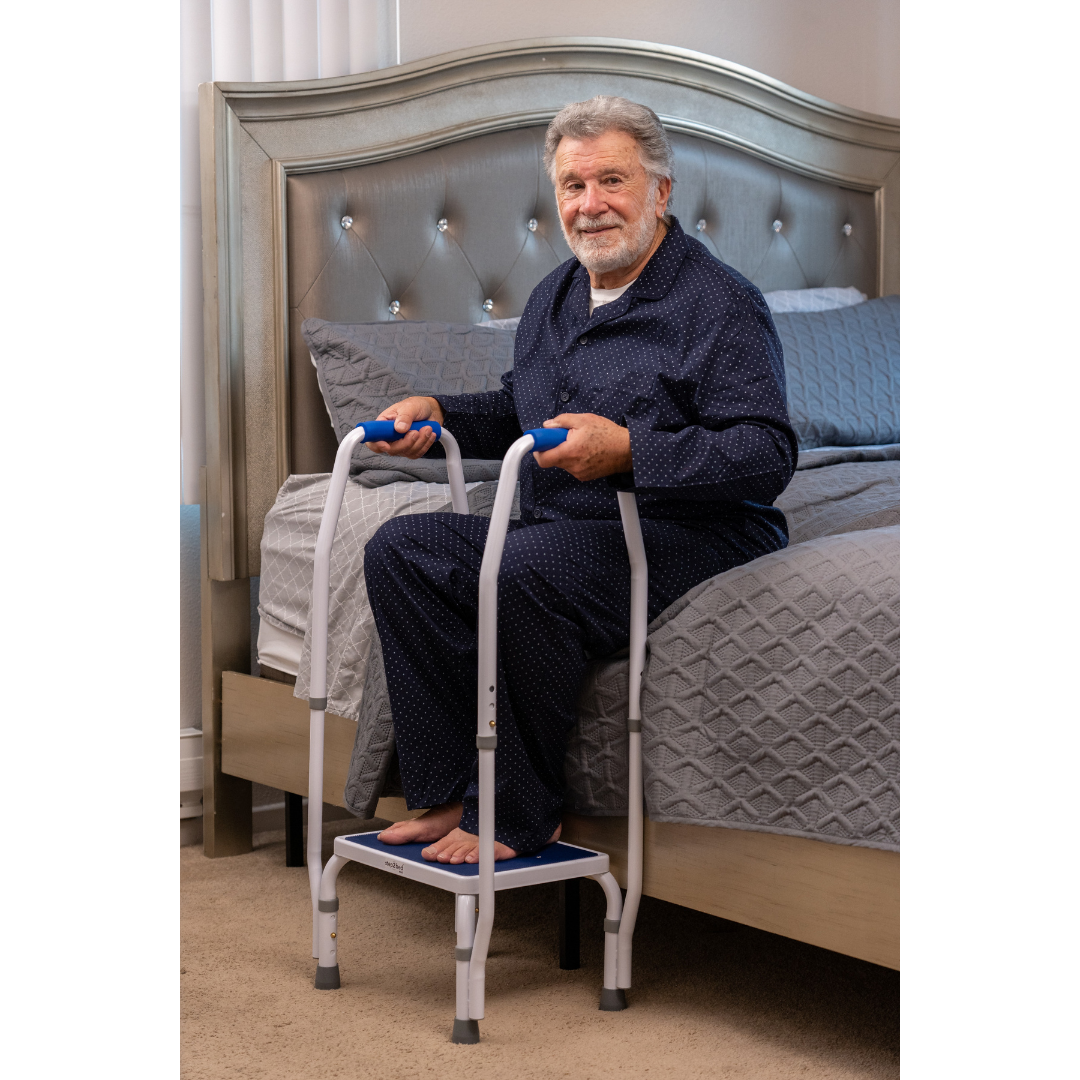 An elderly man in navy pajamas sits on the beds edge, using the Step2Bed Mini Bedside Stepper as a mobility aid, with his feet on its platform, in a carpeted bedroom.