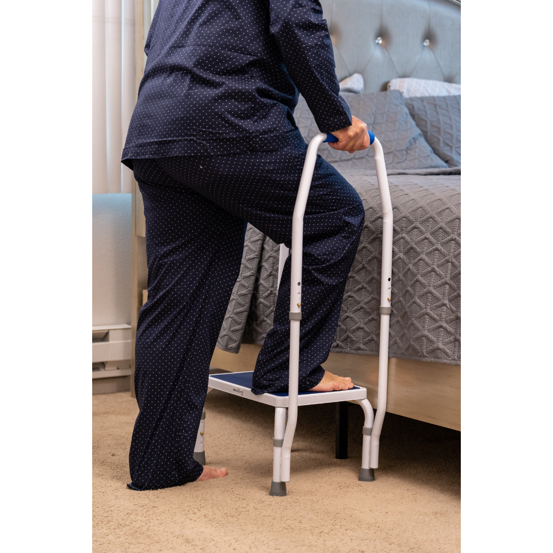 Wearing navy pajamas, a person uses the Step2Bed Mini Bedside Stepper with handles to safely get into a bed with gray bedding in a carpeted bedroom, supporting fall prevention and offering added mobility assistance.