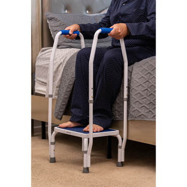 An older adult in pajamas uses the Step2Bed Mini Bedside Stepper with handrails beside the bed, relying on this mobility aid for support and fall prevention when getting in or out of bed.