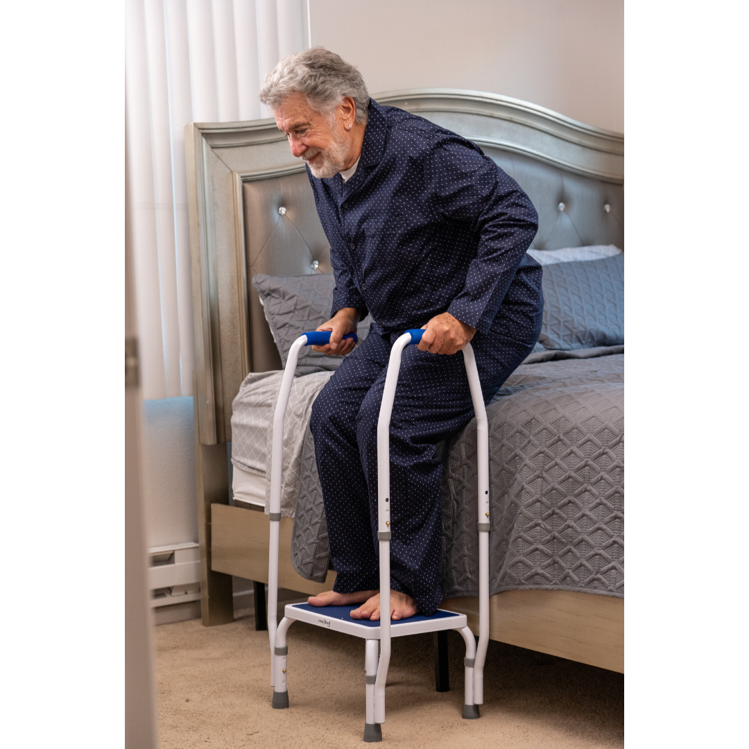 An elderly man in pajamas uses the Step2Bed Mini Bedside Stepper by his bed, gripping the handles with both feet on the platform in a softly lit bedroom, promoting safe and easy standing for fall prevention.