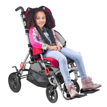 A smiling girl with curly hair sits in a Circle Specialty Strive EZ Adaptive Stroller—red and black, secured with a safety harness—wearing a colorful jacket, light blue jeans, and pink boots against a white background.