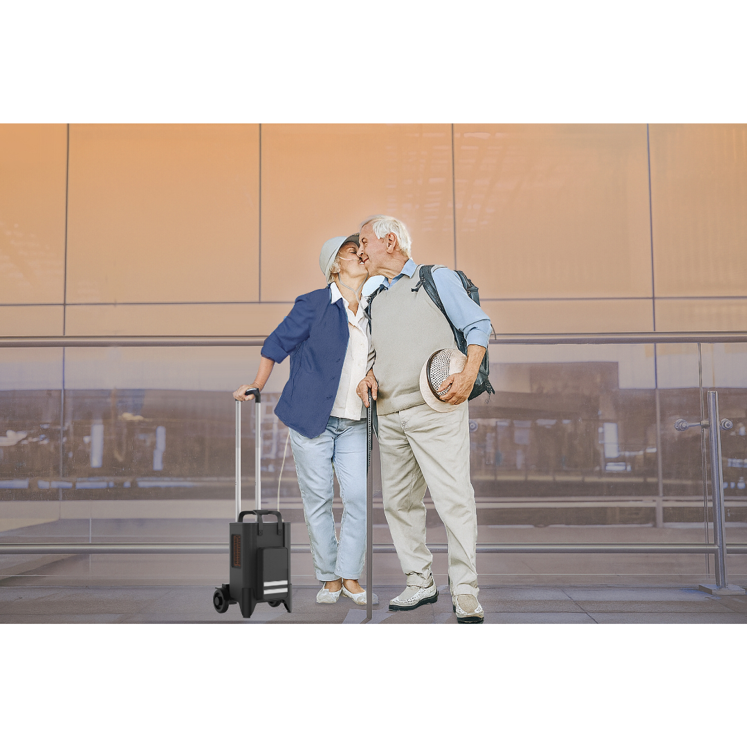 An older couple stands at an airport, sharing a kiss. The woman holds a suitcase, boarding pass, and the Rhythm TOC3 Transportable Oxygen Concentrator; the man carries a backpack and hat. Both wear casual light clothes and glasses.