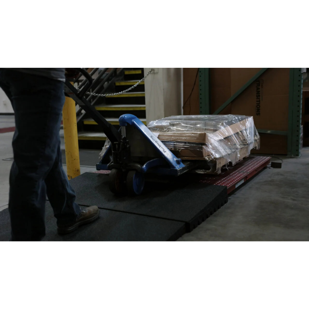 A person moves a shrink-wrapped pallet up an EZ-Access TRANSITIONS® Modular Entry Mat with a blue pallet jack in a dimly lit warehouse, ensuring safe passage. Only their legs are visible.