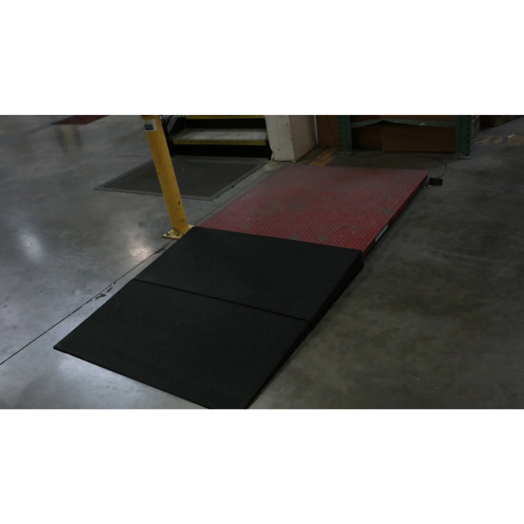 An EZ-Access TRANSITIONS® Modular Entry Mat and a black portable wheelchair ramp are set on a concrete floor, providing a safe transition to a slightly raised red platform in an indoor industrial space.