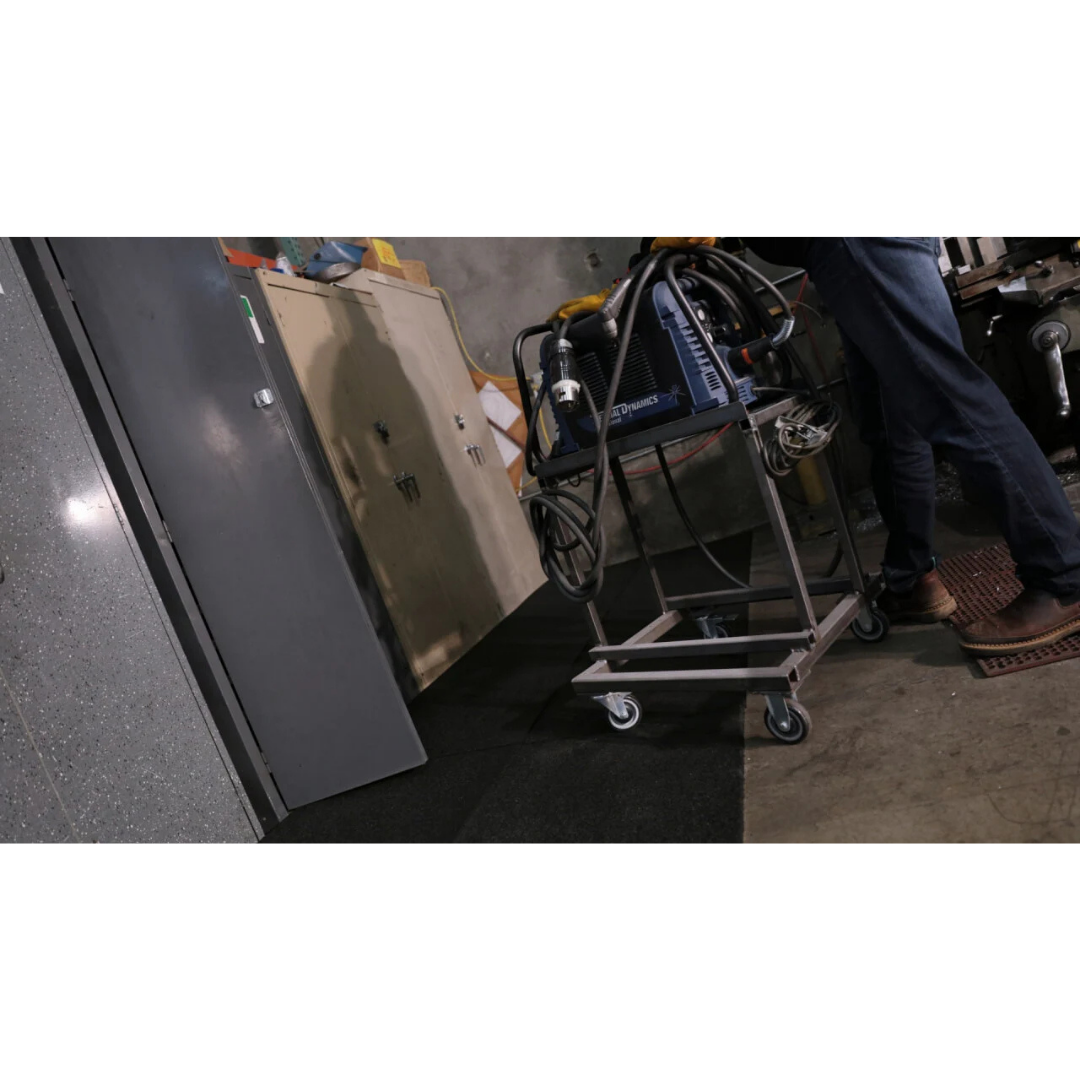 A person stands by a metal cabinet and a wheeled cart with a welding machine in a workshop with concrete floors and walls, featuring the EZ-Access TRANSITIONS® Modular Entry Mat for an eco-friendly, safe transition at the entryway.