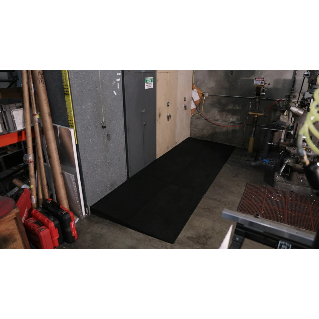 An EZ-Access TRANSITIONS® Modular Entry Mat is placed on the floor before gray metal cabinets and lockers in an industrial room, offering a safe transition near exposed pipes, wires, and equipment.