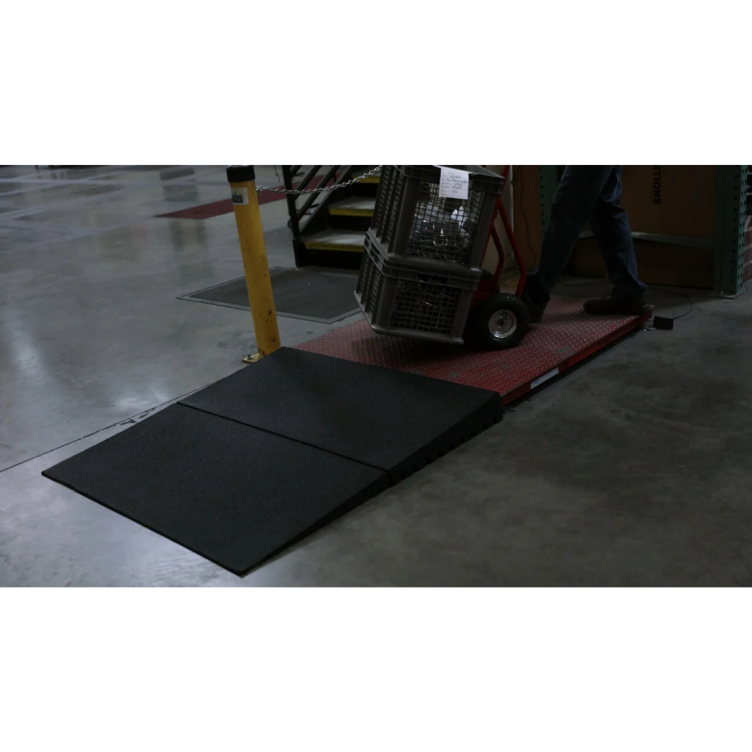 A person uses a hand truck to move a bin up a black portable ramp and over EZ-Access TRANSITIONS® Modular Entry Mats onto a red platform in an industrial warehouse.