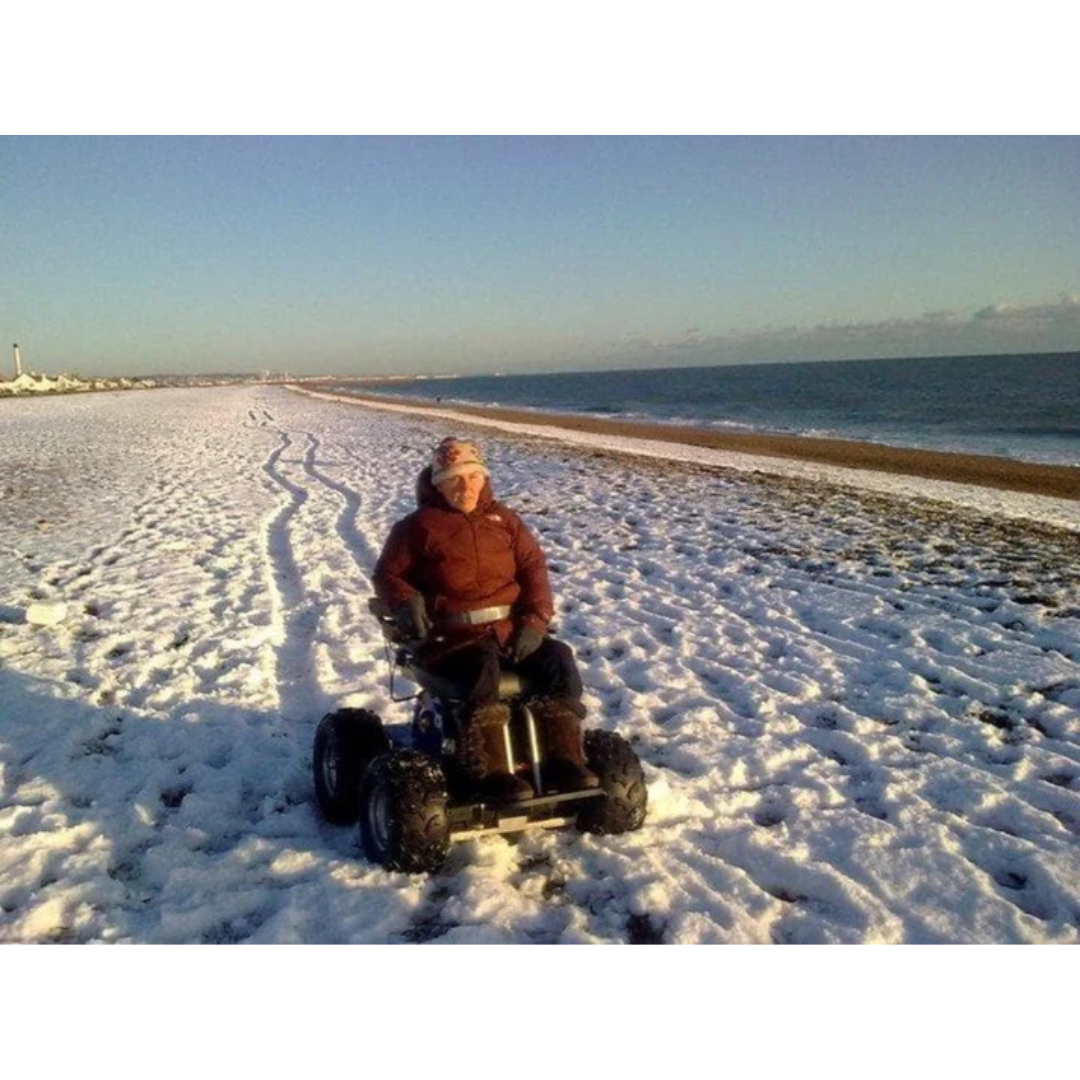 A person in a wheelchair with large off-road wheels travels along a snowy beach near the shore in a Wheelchair88 All-Terrain Stair Climbing 4x4 Recreational Electric Vehicle, leaving tracks in the snow with the sea and blue sky behind them.