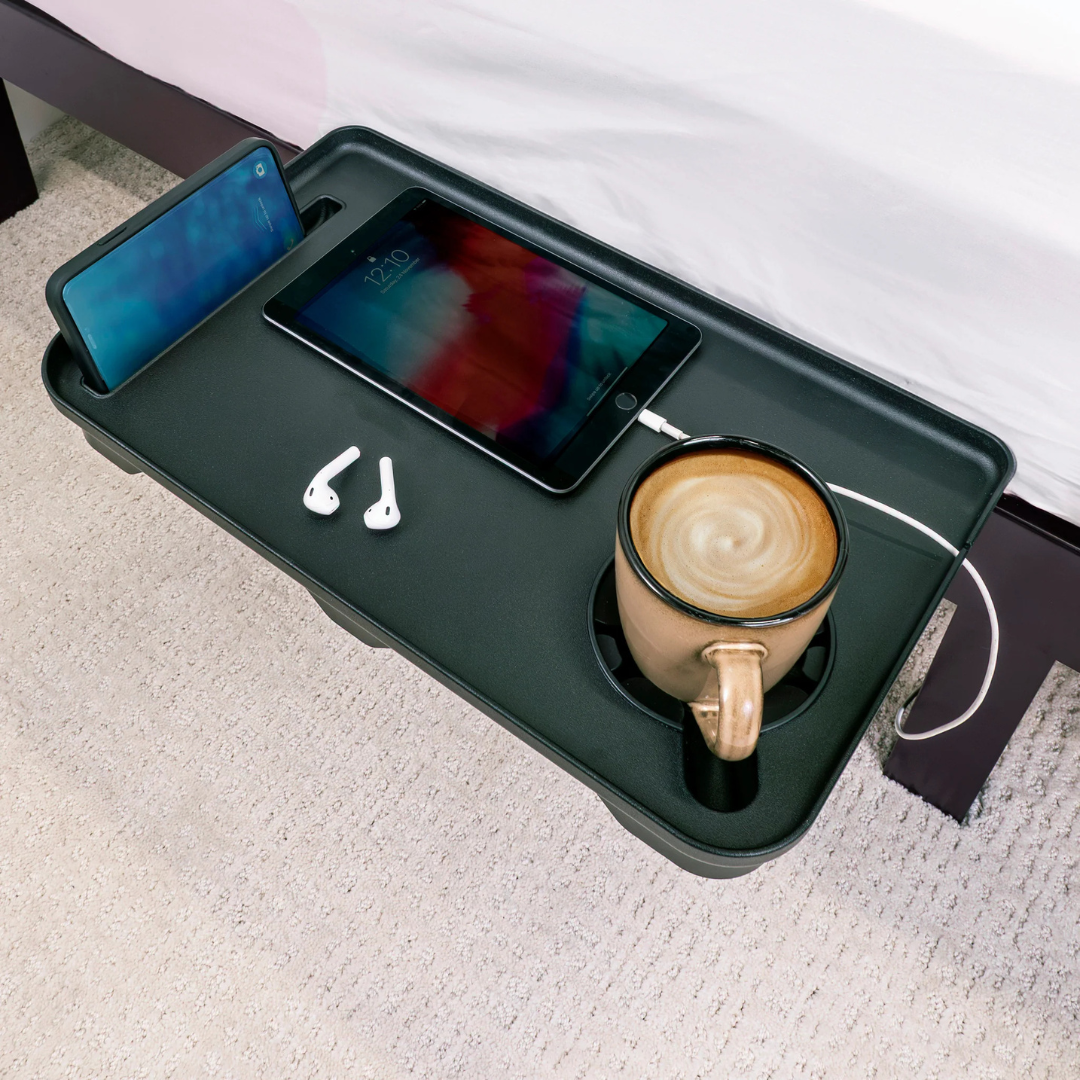 The Stander Wonder Bedside Shelf Tray is a black, space-saving shelf that neatly holds and charges your smartphone, tablet, wireless earbuds, and a coffee mug beside your bed on a light carpeted floor.