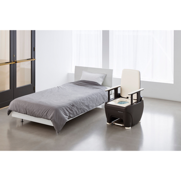 A minimalist bedroom with a low bed in gray bedding and the Wrappon® ORB Portable Commode Toilet with auto waste sealing, featuring built-in controls, in a bright room with large windows and a glass door.