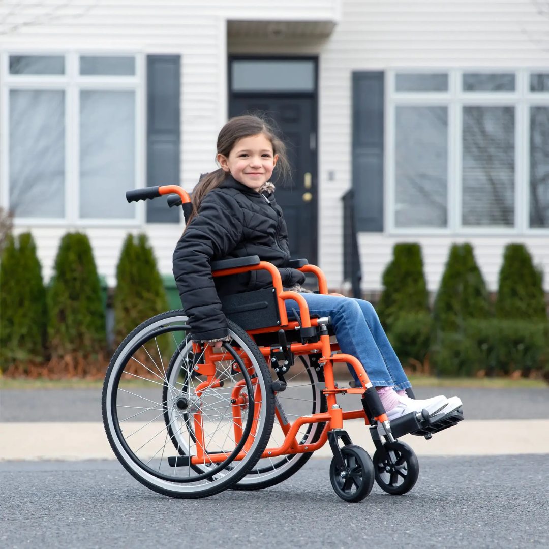 A young girl in a black jacket and jeans smiles while sitting in a bright orange Circle Specialty ZIGGO Pediatric Wheelchair outside in front of a house with large windows and shrubs.