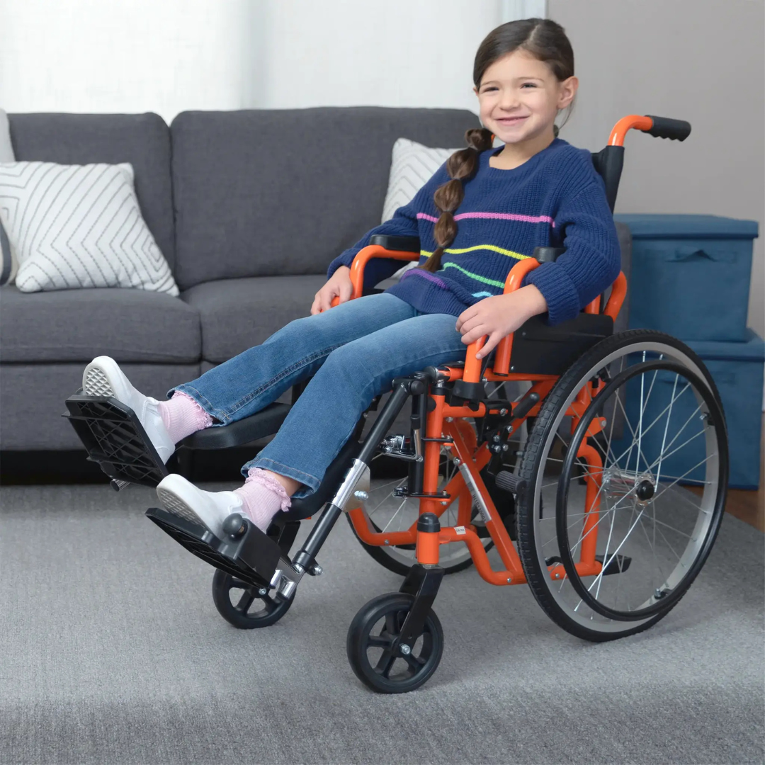 A young girl with a long brown braid, in a blue sweater and jeans, smiles while sitting in a Circle Specialty ZIGGO Pediatric Wheelchair in an orange color, with a gray couch and cushions visible in the living room background.