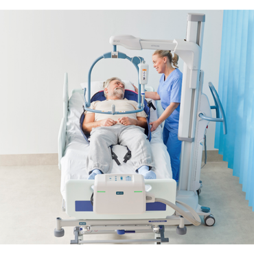 An older man rests in a hospital bed with the ARJO Maxi Move 5 Multifunctional Patient Floor Lift above him, while a nurse in blue scrubs stands beside the bed, smiling and assisting him in a bright, clinical room.