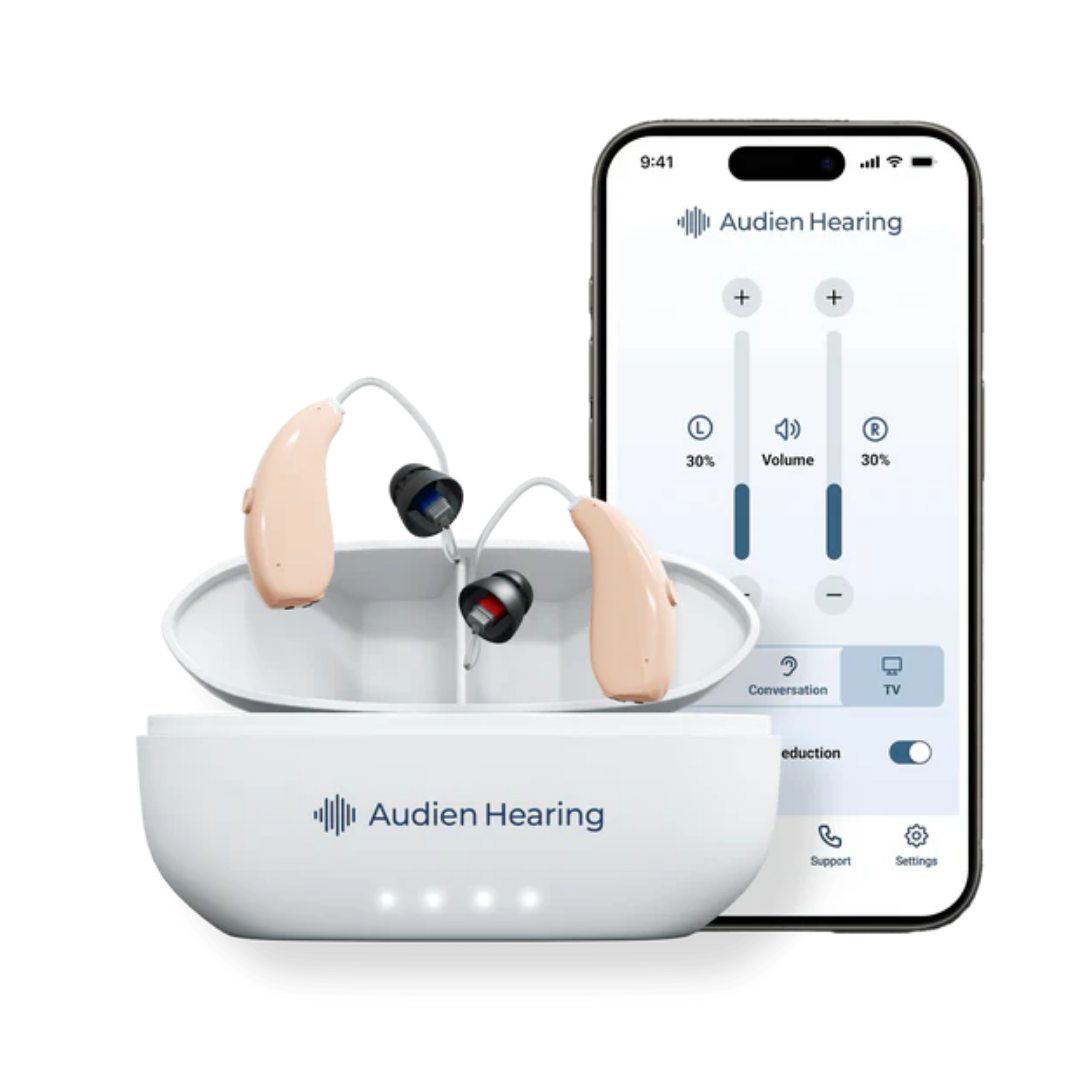A pair of beige Audien Hearing ION Pro Over-The-Ear High Def Hearing Aids rest in an open white charging case, with a smartphone behind them displaying the Audien Hearing app for streaming and personalized settings.