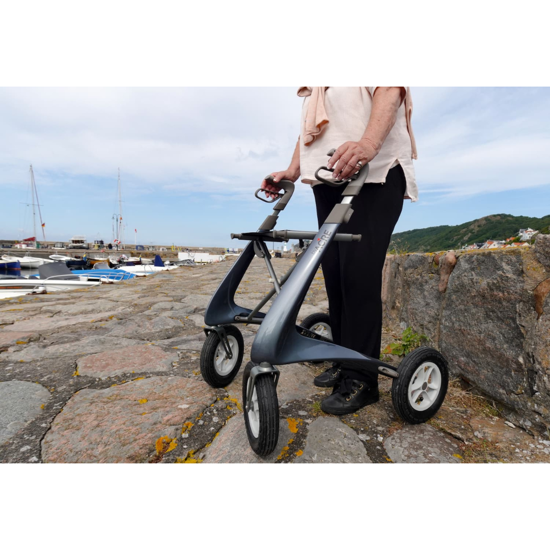 An older person stands outdoors on a stone path with their ByACRE Carbon Overland All-Terrain Rollator—perfect for adventurous walks. Boats and water appear in the background, with lush greenery and a hillside to the right.