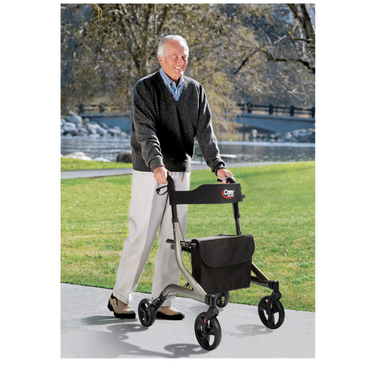 An older man smiles as he walks outside on a sidewalk using the Carex Crosstour Rolling Walker with Euro Design. He wears a dark sweater over a collared shirt and light pants. A park with grass, trees, and a bridge is in the background.