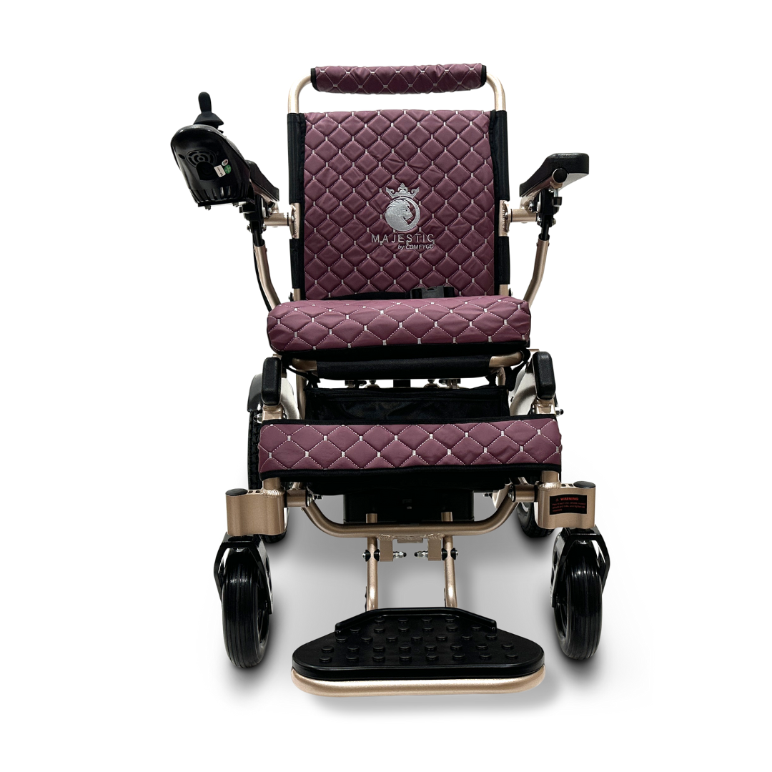 Front view of the ComfyGo MAJESTIC IQ-8000, a travel-friendly electric wheelchair featuring a quilted burgundy seat and backrest. It includes armrests, joystick control, and a footrest. The lightweight design ensures ease of use, with the Majestic brand elegantly embroidered on the backrest.