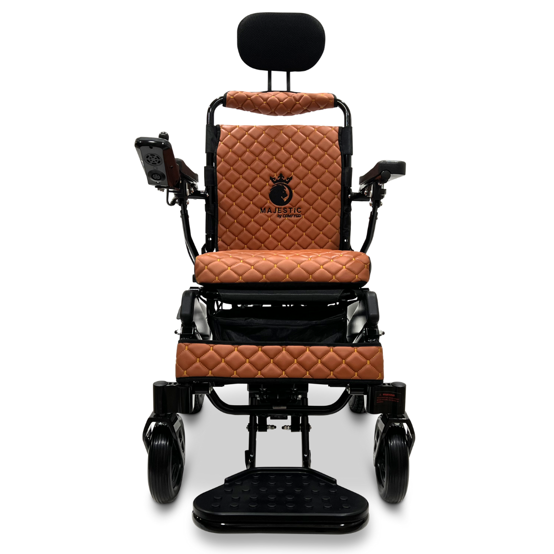 A front view of the ComfyGo MAJESTIC IQ-9000 Auto Recline Remote Controlled Electric Wheelchair highlights a plush, quilted brown leather seat and backrest. It includes a headrest, armrests with integrated controls, and a footplate. The Majestic logo is visible on the backrest, enhanced by brushless silent motors for a smooth ride.