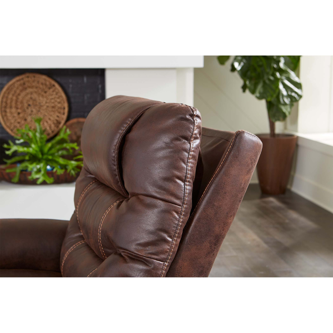 Close-up of the headrest and upper back of the Golden Tech PR446 DeLuna™ Dione Recliner with Lift Assist in brown faux leather, shown in a living room with a softly blurred plant and fireplace in the background.