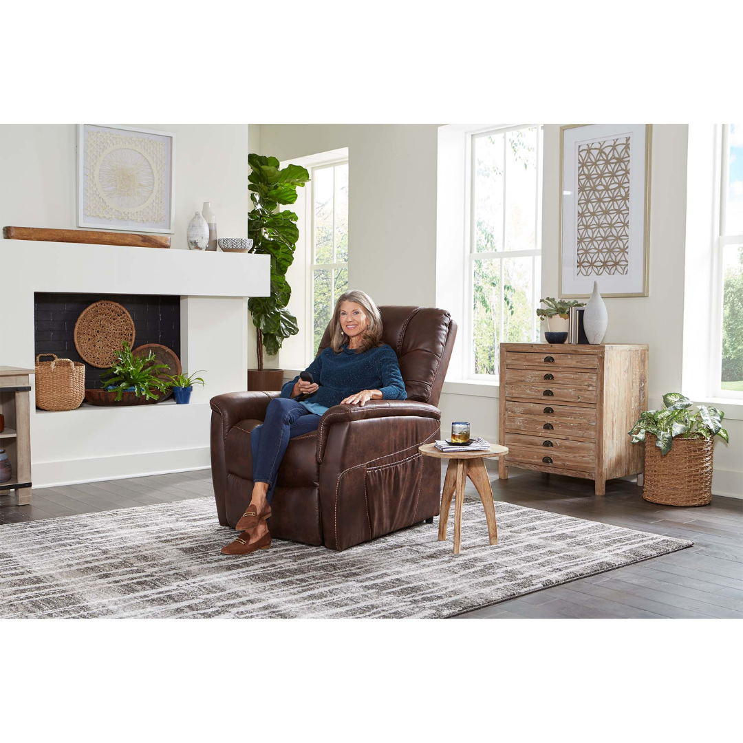 A woman smiles, holding a remote, as she relaxes in a Golden Tech PR446 DeLuna™ Dione Recliner with Lift Assist in a bright living room featuring large windows, modern decor, plants, and a gray patterned rug.