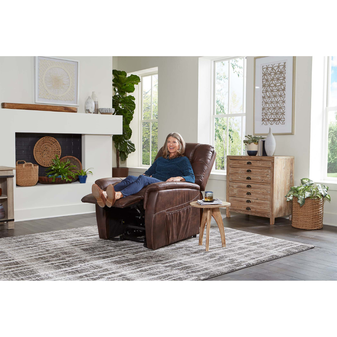 A woman relaxes with her legs up in a bright, modern living room on the Golden Tech PR446 DeLuna™ Dione Recliner with Lift Assist, surrounded by large windows, wooden furniture, plants, and a patterned rug.