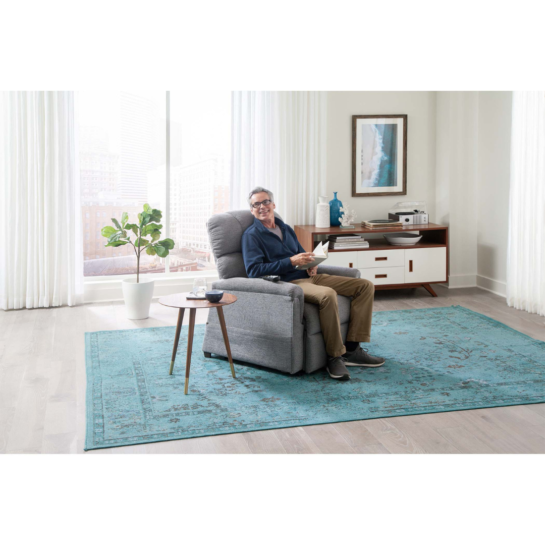 A smiling older man relaxes in the Golden PR531 Comforter Recliner w/ Lift Assist, holding a remote and a book in a bright living room with a turquoise rug, side table, potted plant, and white sideboard, enjoying customized comfort.