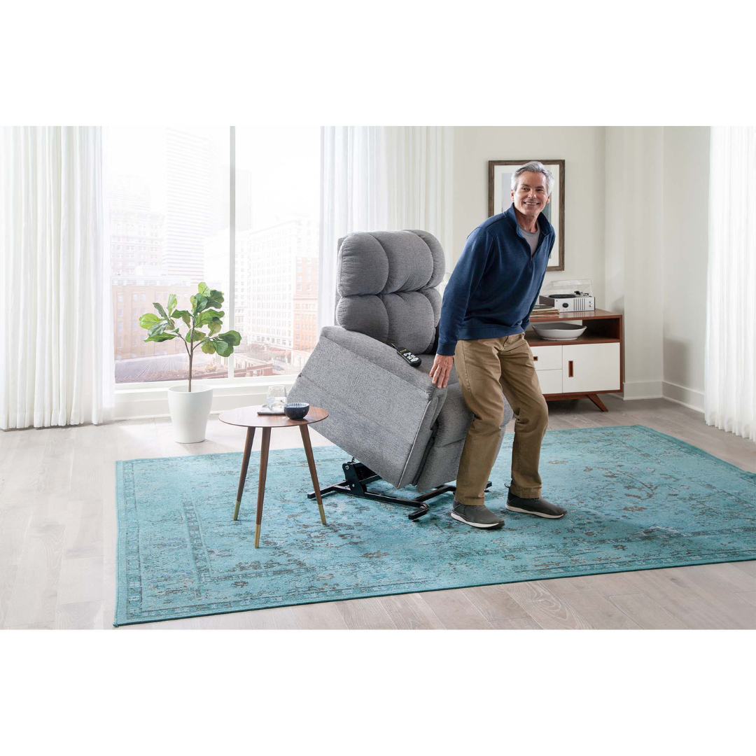 A person leans against a Golden PR535 MaxiComfort Recliner w/ Lift Assist - Medium, partially raised for ergonomic support, smiling in a blue sweater and khaki pants. Nearby, a side table with a smartphone and remote complements the inviting ambience of the room with its large window, potted plant, and blue rug.