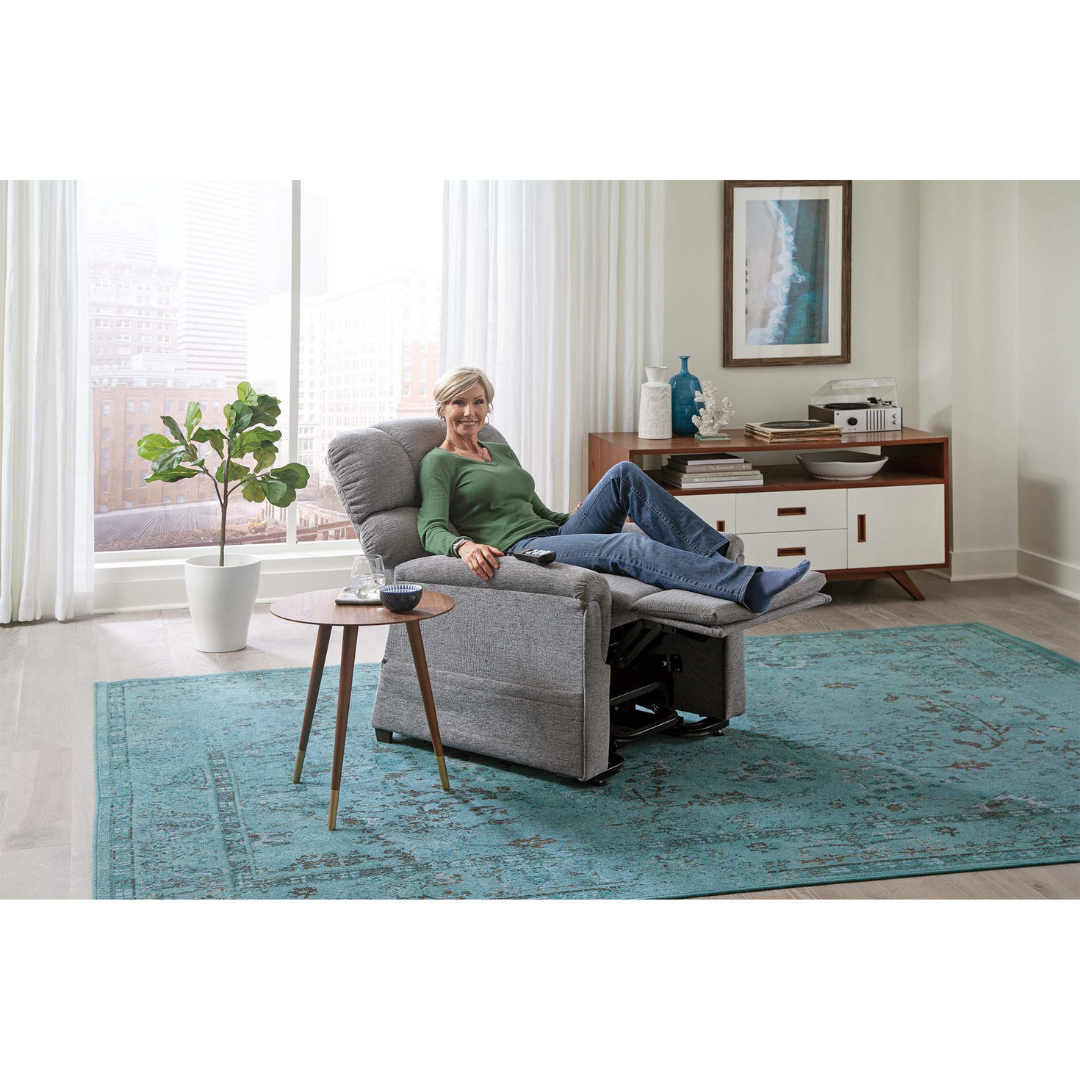 A woman relaxes and smiles in the Golden Tech PR535 MaxiComfort Recliner w/ Lift Assist, her feet up in a bright living room with large windows, a blue rug, potted plant, and wooden sideboard in the background.