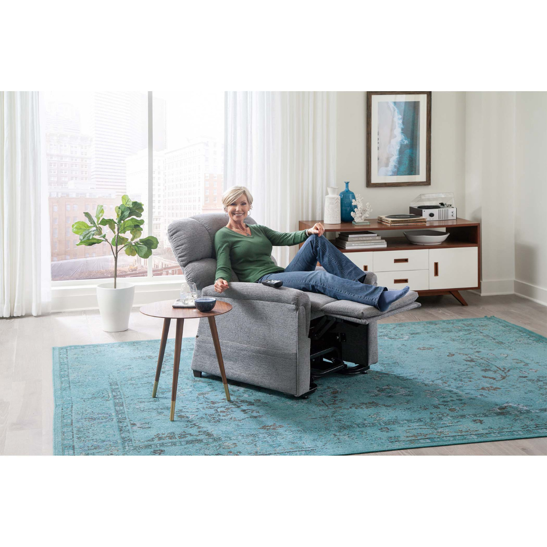A woman relaxes in a Golden PR531 Comforter Recliner w/ Lift Assist, smiling with her feet up and holding the remote. The inviting room features large windows, a plant, blue rug, side table, and console, offering personalized comfort.
