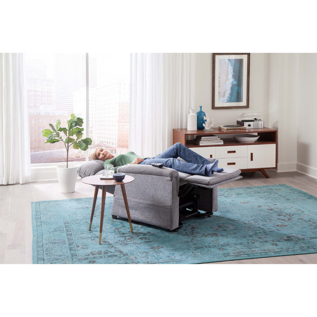 A woman relaxes with her feet up on a gray Golden PR531 Comforter Recliner w/ Lift Assist in a bright living room, featuring a blue rug, round table, potted plant, sideboard, and large windows with city views and sheer curtains.