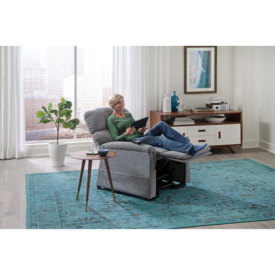 A woman relaxes with custom comfort on the Golden PR531 Comforter Recliner w/ Lift Assist, reading a tablet in a bright, modern living room featuring large windows, a blue rug, a potted plant, and mid-century furniture.