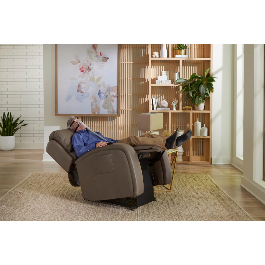 A person in a blue shirt and brown pants is lounging in a Golden Tech PR761 EZ Sleeper Recliner with Lift Assist & Twilight Recline. They appear relaxed, using an eye massage device, surrounded by plants, vases on shelves, and abstract wall art in the modern living room.
