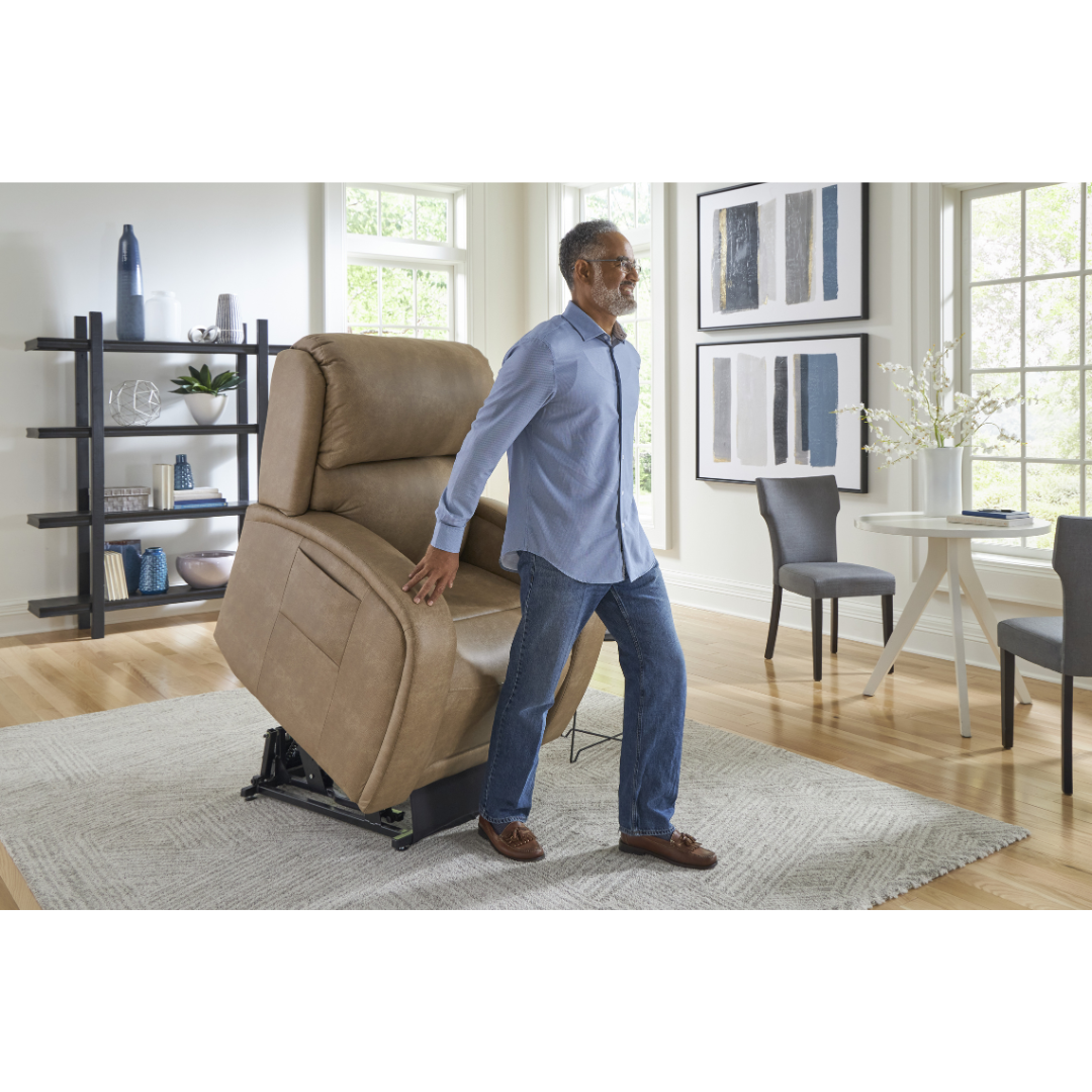 An elderly man stands supported by a Golden Tech PR761 EZ Sleeper Recliner with Lift Assist & Twilight Recline in a bright living room. He wears glasses, a light blue shirt, and jeans. The room features large windows, modern art, and a round table with chairs on a patterned rug.