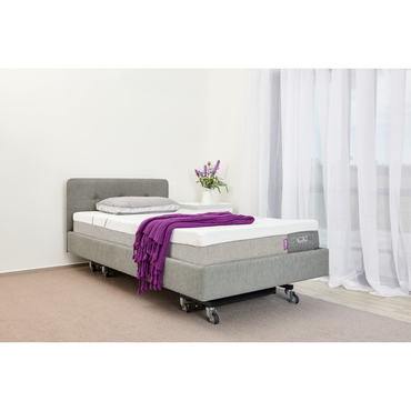 The iCare Medical IC100 Static Partner Non-Adjustable Bed in gray features iCare bed rails and wheels, with a white mattress, gray pillow, and purple blanket. A small white table with flowers stands beside it in a bright room with sheer curtains.